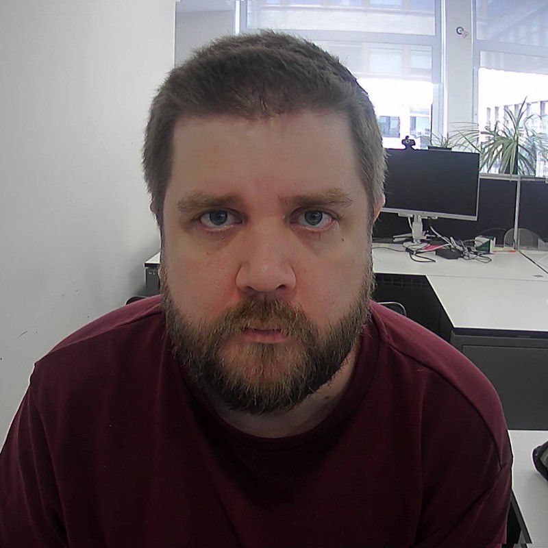 White guy with glasses and a beard, not smiling which is explained below